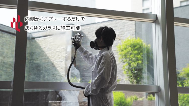 防護服を着た作業者が、室内側から窓ガラスにスプレーガンでコーティングを吹き付けている様子